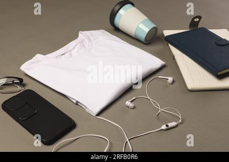 Flache Lagen aus weißem T-Shirt, Smartphone, Ohrhörern und Notizbüchern mit Kopierbereich auf grauem Hintergrund Stockfoto