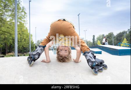 Der junge süße Junge, der Spaß auf Rollschuhen hat, steht kopfüber auf, lässt sich verwöhnen, während er an einem Sommertag im Park das Schlittschuhlaufen lernt. Kinder und Sport, Hobb Stockfoto