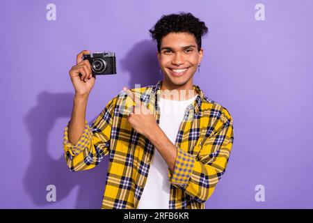 Foto eines coolen, positiven Mannes im karierten gelben Hemd mit Finger-Vintage-Fotokamera, isoliertem lilafarbenen Hintergrund Stockfoto