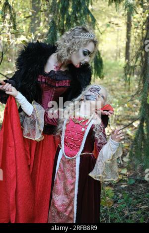 Eine Frau in Form eines Vampirs oder einer Zauberin posiert im Wald mit einem Mädchen in märchenhaftem Make-up und mittelalterlichem Kleid. Halten Sie Ausschau nach Halloween. Vertikal Stockfoto