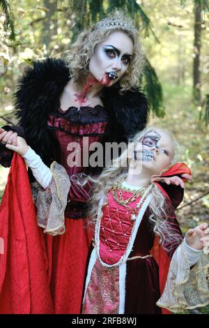 Eine Frau in Form eines Vampirs oder einer Zauberin posiert im Wald mit einem Mädchen in märchenhaftem Make-up und mittelalterlichem Kleid. Bild für Halloween. Vertikal Stockfoto