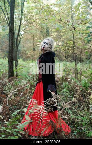 Eine Frau im Bild eines Vampirs oder einer Hexe mit weißer Schminke und in einem mittelalterlichen Kleid posiert im Wald vor der Kamera. Vertikales Foto Stockfoto