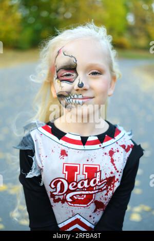 Ein Mädchen, gekleidet als Cheerleaderin und mit Make-up auf der Hälfte ihres Gesichts, in Form eines Skeletts, lächelt und schaut in die Kamera. Vertikales Foto Stockfoto