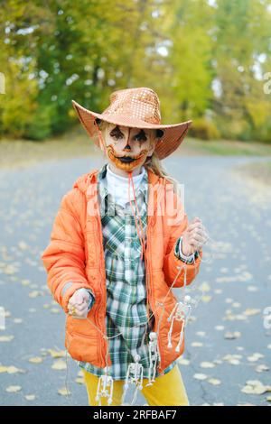 Ein Mädchen mit orangefarbenem Cowboyhut und Make-up im Gesicht posiert für die Kamera mit einer Dekoration von Skeletten. Halloween-Feier. Vertikal Stockfoto