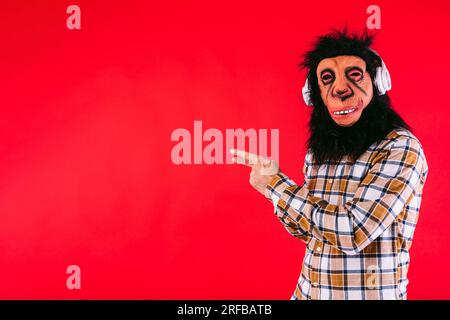 Mann mit Schimpansen-Affenmaske und kariertem Hemd, trägt Kopfhörer, zeigt mit dem Finger auf den Kopierbereich, auf rotem Hintergrund. Stockfoto