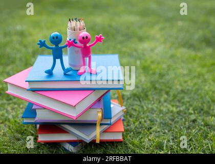 Ein Stapel bunter Bücher liegt auf dem Gras, auf dem zwei lustige, pinkfarbene und blaue Figuren als Souvenir, ein Satz Bleistifte. Karte für Lehrer. Zurück in die Schule. Konz Stockfoto