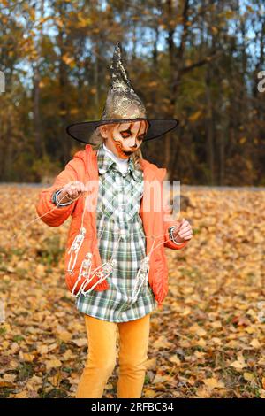 Ein Mädchen in einer orangefarbenen Jacke, einem Hexenhut und mit Make-up im Gesicht hält eine Dekoration von Skeletten in ihren Händen. Halloween-Feier. Verti Stockfoto