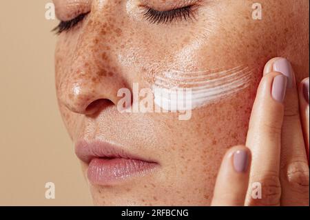 Nahaufnahme des weiblichen Gesichts mit Sommersprossen. Detailgetreue Aufnahme einer Frau, die Feuchtigkeitscreme aufträgt. Stockfoto