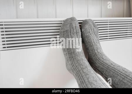 Beine in Stricksocken, die sich über dem elektrischen Heizer erwärmen. Eine gefrorene Frau, die warme Wollsocken trägt, eiskalt vor der Winterkälte. Unbehagen, Zeit in der Wohnung zu verbringen Stockfoto