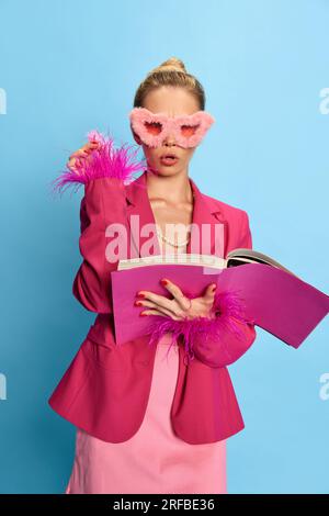 Porträt eines charmanten Mädchens, eine Frau mit Glamour und ungewöhnlichen Brillen, die ein Lehrbuch in der Hand hält und auf hellblauen Studiohintergrund in die Kamera schaut. Mode. Stockfoto