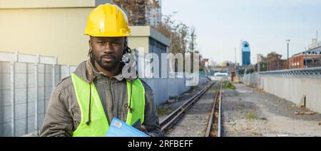 Banner eines erwachsenen männlichen Bauherstellers afrikanischer ethnischer Herkunft, trägt einen gelben Sicherheitshelm und eine reflektierende Weste, steht auf Eisenbahnschienen und hält blaue Ordner und Stockfoto