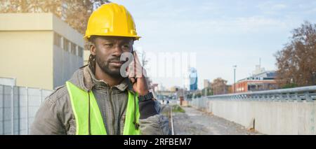 Banner eines Bauarbeiters afrikanischer ethnischer Herkunft mit gelbem Helm und reflektierender Weste, der am Telefon telefoniert und draußen auf den Gleisen steht. Stockfoto