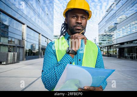 Porträt eines schwarzen Mannes mit Bart und Dreadlocks, Architekt in gelbem Sicherheitshelm und Weste, der draußen steht und denkt, einen Ordner zu halten, Kopierraum. Stockfoto