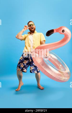 Fröhlicher und entspannter Mann mit Bart in Badehosen und gelbem Hemd mit Schwimmkreis vor blauem Studiohintergrund. Sommerferien Stockfoto