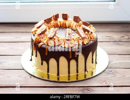 Erdnussbutter-Creme-Kuchen mit geschmolzener Schokolade, Schokoriegeln, Erdnussbissen und gesalzenem Karamell auf Holzhintergrund Stockfoto