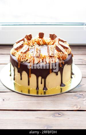 Erdnussbutter-Creme-Kuchen mit geschmolzener Schokolade, Schokoriegeln, Erdnussbissen und gesalzenem Karamell auf Holzhintergrund Stockfoto