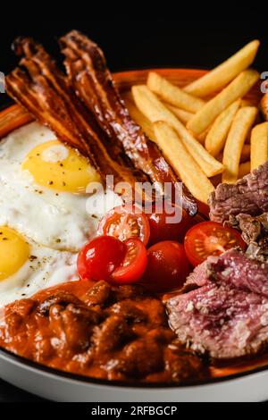 Traditionelles englisches Frühstück. Spiegeleier mit Speck, Tomaten, Bohnen, pommes auf schwarzem Hintergrund. Nahaufnahme. Stockfoto