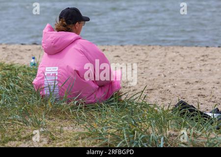 Frau in pinkfarbener Jacke, die im Juli in Sandbanks, Poole, Dorset UK am Strand sitzt Stockfoto