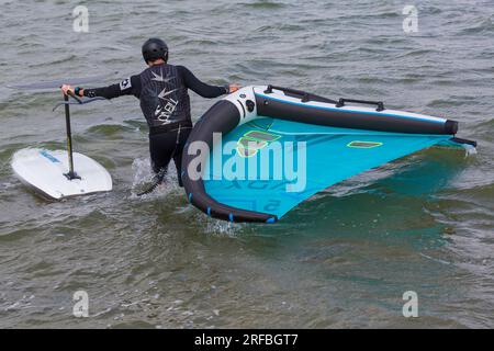 Ein Mann trägt einen O'Neill Neoprenanzug und trägt Flügelfolien ins Meer, bereit für Flügelfolien im Juli in Sandbanks, Poole, Dorset, Großbritannien Stockfoto