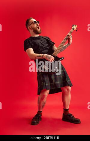 Bärtiger Mann in Sonnenbrille und Rock, Kilt spielt Gitarre vor rotem Studiohintergrund. Rock 'n' Roll Stockfoto