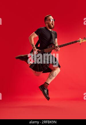 Bärtiger Mann in Sonnenbrille und Rock, Kilt spielt Gitarre vor rotem Studiohintergrund. Emotionaler Musiker Stockfoto