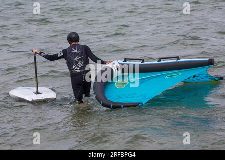 Ein Mann trägt einen O'Neill Neoprenanzug und trägt Flügelfolien ins Meer, bereit für Flügelfolien im Juli in Sandbanks, Poole, Dorset, Großbritannien Stockfoto