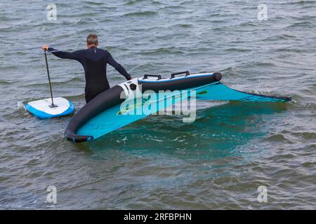 Ein Mann trägt einen O'Neill Neoprenanzug und trägt Flügelfolien ins Meer, bereit für Flügelfolien im Juli in Sandbanks, Poole, Dorset, Großbritannien Stockfoto