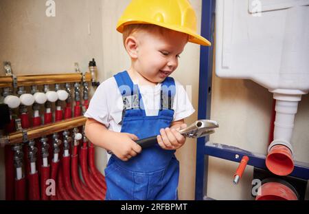 Der kleine Junge sieht sich das Bauwerkzeug an und lächelt, während er neben der Installation von Haushaltsrohren steht. Kind im Schutzhelm und Arbeitsanzug mit Reparaturinstrumentschlüssel. Hausrenovierungskonzept. Stockfoto
