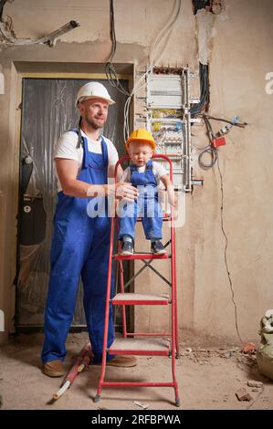 Ein kleiner Junge sitzt auf der Leiter und hält das Bauwerkzeug, während der Mann neben dem Kind steht. Männlicher Arbeiter und Kind, das sich in der Nähe einer Schalttafel in einer Wohnung befindet, die gerade renoviert wird. Stockfoto