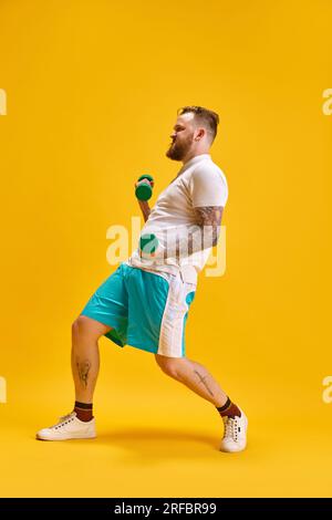 Durchgehendes Bild eines bärtigen Mannes im Sportbekleidungstraining mit Kurzhanteln vor gelbem Studiohintergrund. MEME-Workout Stockfoto