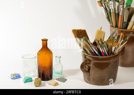 Künstlerische Pinsel, farbenfrohe Steine, alte Flaschen und Malwerkzeuge in alten Keramiktöpfen auf weißem Hintergrund. Künstlerische Komposition. Stockfoto