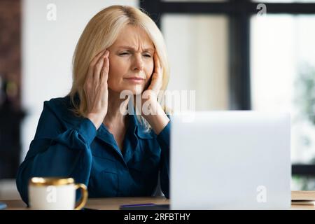 Gestresste, reife Geschäftsfrau, die bei der Arbeit an einem Notebook Kopfschmerzen hat, Frau, die auf den Bildschirm schaut und Bügel reibt Stockfoto