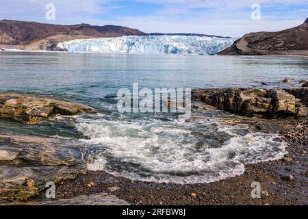 Meereslandschaft einer Welle, die sanft über niedrige Felsen bricht, mit dem massiven eqip-Sermia-Gletscher im Hintergrund Stockfoto