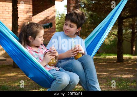 Süße kaukasische Schulkinder, die Smoothies trinken und sich in der Hängematte im Hinterhof entspannen. Kinder. Freizeit. Lifestyle Stockfoto
