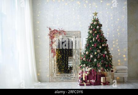 Gemütliche weihnachtseinrichtung des Wohnzimmers mit kunstvollem weihnachtsbaum und großem Spiegel in der Nähe des Fensters. Stockfoto