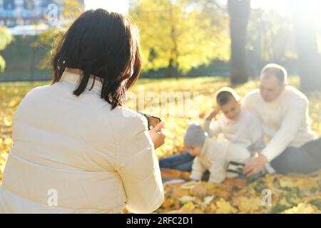 Eine glückliche vierköpfige Familie verbringt Zeit im Herbstpark. Mutter macht Fotos von ihrer Familie oder nimmt Videos auf dem Mobiltelefon auf. Horizontales Foto Stockfoto