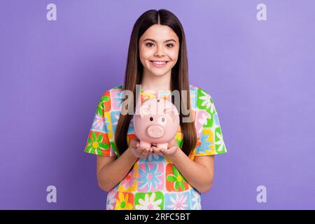 Foto einer attraktiven Teenagerin mit Sparschwein und stilvoller Kleidung mit Blumenmuster, isoliert auf lila Hintergrund Stockfoto