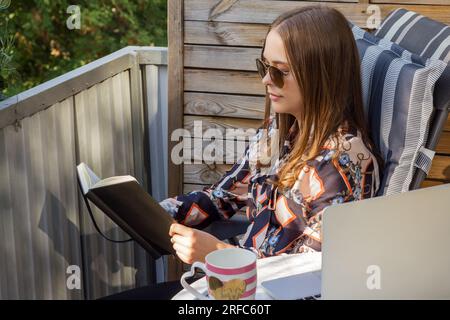 Eine junge Frau, die auf dem Balkon sitzt und ein Buch oder Notizbuch liest. Outdoor-Szene. Stockfoto