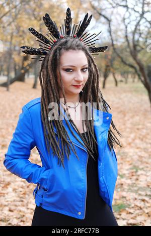 Eine Frau mittleren Alters in Dreadlocks und ein Reifen auf ihrem Haar in Federn wandert im Herbstpark. Die Frau schaut in die Kamera. Vertikales Foto Stockfoto