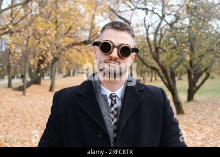 Ein Mann in einem Business-Anzug, Mantel und Steampunk-Brille posiert in einem Herbstpark. Horizontales Foto Stockfoto