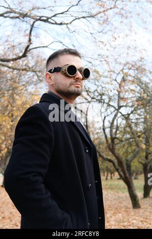 Ein Mann in einem Business-Anzug, Mantel und Steampunk-Brille posiert in einem Herbstpark. Vertikales Foto Stockfoto