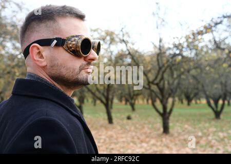 Ein Mann in einem Business-Anzug, Mantel und Steampunk-Brille posiert in einem Herbstpark. Horizontales Foto Stockfoto