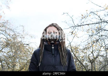 Porträt einer Frau in einer schwarzen Jacke mit Dreadlocks und einer Gasmaske mit Stacheln. Eine Frau, die im Herbstpark posiert. Horizontales Foto Stockfoto