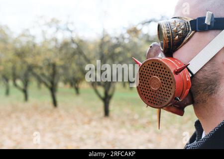 Ein Mann mit schwarzem Mantel, Business-Anzug, Gasmaske und Steampunk-Schutzbrille posiert in einem Herbstpark. Horizontales Foto Stockfoto