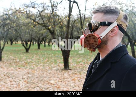 Ein Mann mit schwarzem Mantel, Business-Anzug, Gasmaske und Steampunk-Schutzbrille posiert in einem Herbstpark. Horizontales Foto Stockfoto