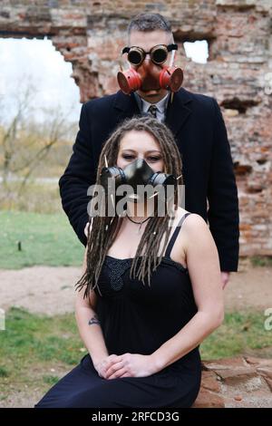 Zwei Personen in schwarzer Kleidung und Gasmasken posieren vor dem Hintergrund einer zerstörten Ziegelmauer. Steampunk-Brille auf einen Mann. Vertikales Foto Stockfoto