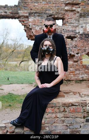 Zwei Personen in schwarzer Kleidung und Gasmasken posieren vor dem Hintergrund einer zerstörten Ziegelmauer. Steampunk-Brille auf einen Mann. Vertikales Foto Stockfoto