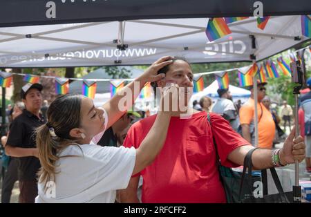 Beim Transfest macht eine Transfrau ein Selfies, während ihr die Transflagge auf die Wange gemalt wird. In Jackson Heights, Queens, New York. Stockfoto