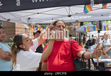 Beim Transfest macht eine Transfrau ein Selfies, während ihr der Transfalg auf die Wange gemalt wird. In Jackson Heights, Queens, New York. Stockfoto