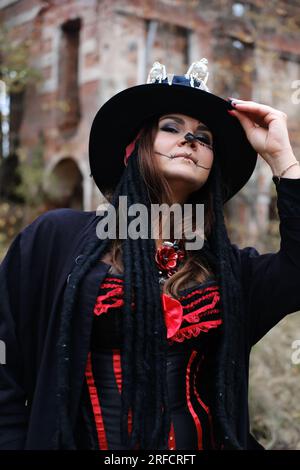 Eine Frau im Bild von Baron Saturday posiert vor dem Hintergrund eines alten Backsteingebäudes. Das Model trägt ein Korsett, eine Strickjacke und einen Hut Stockfoto